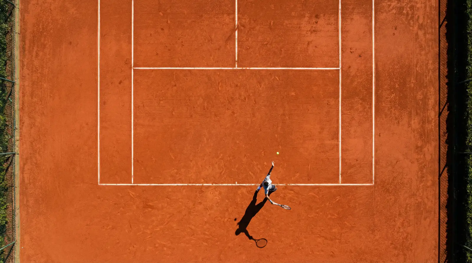 Vista de cima de um court de ténis em terra batida com jogador a preparar o serviço